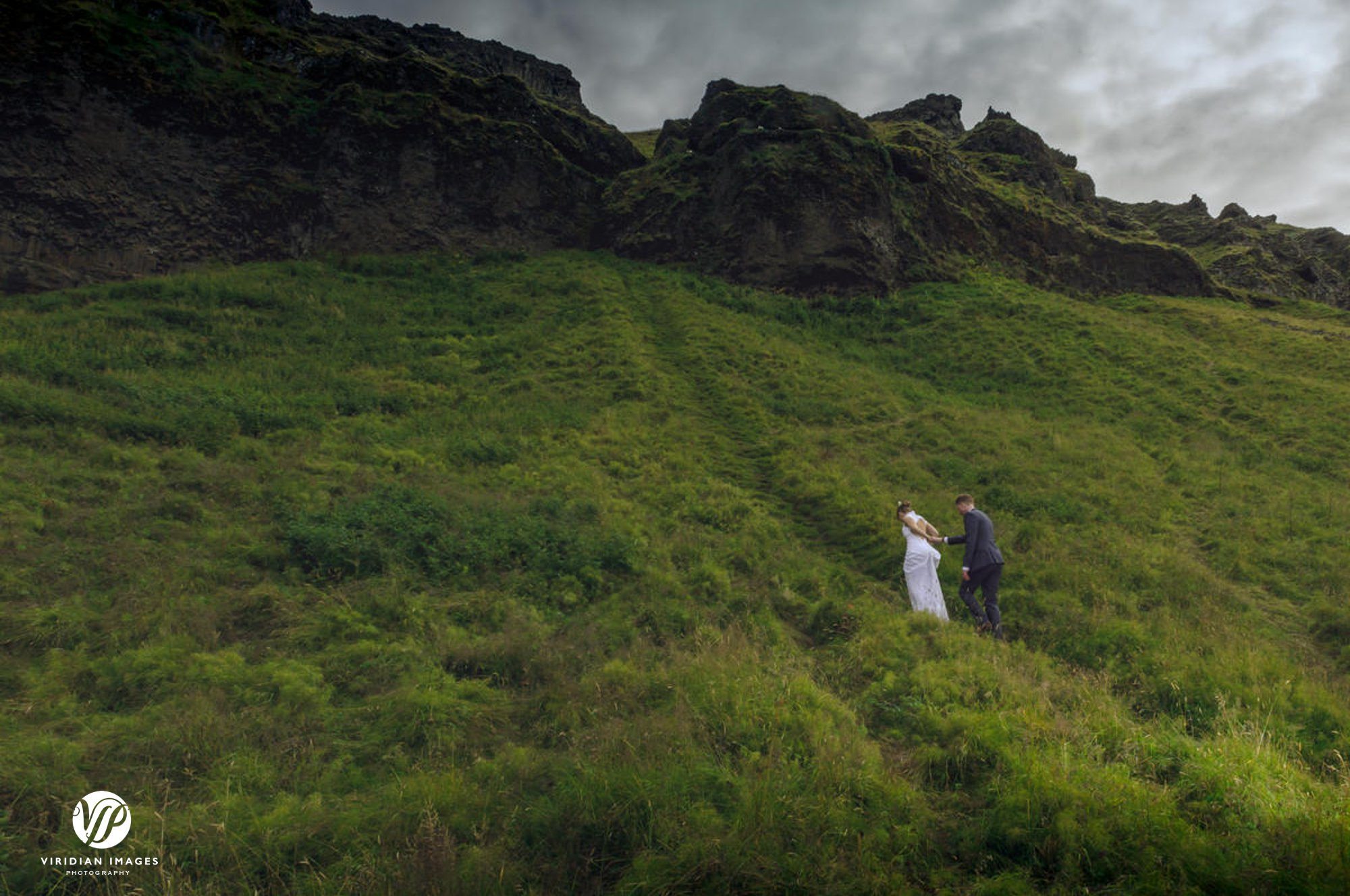 Bride and groom climbing side of mountain by Seljalandsfoss waterfall Iceland
