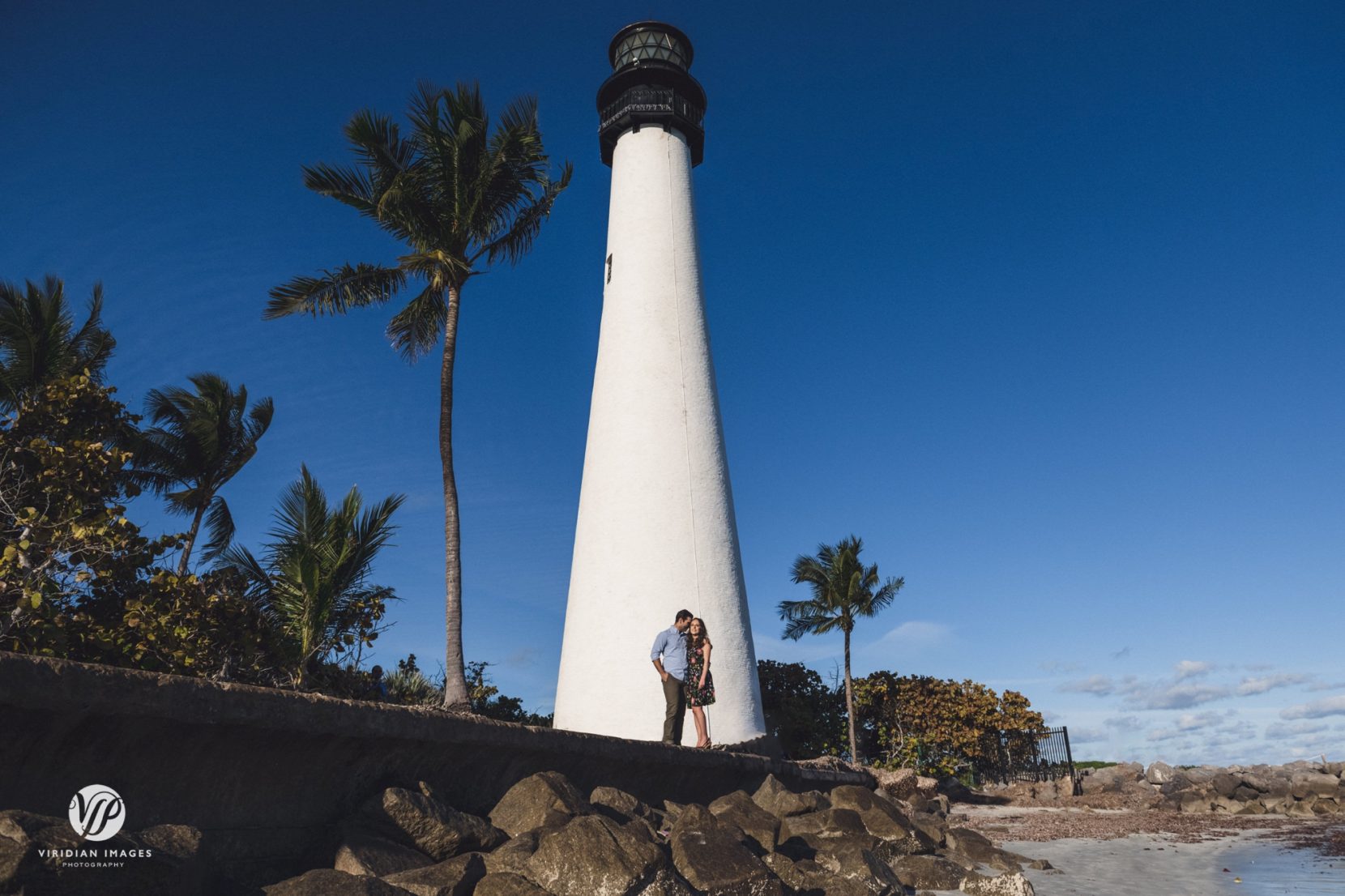 wide shot of couple by beach and lighthouse against bright blue sky