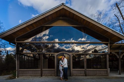 Engagement photo of couple standing in front of reflective windows showing a dramatic sky at Rocky Lake Estate.