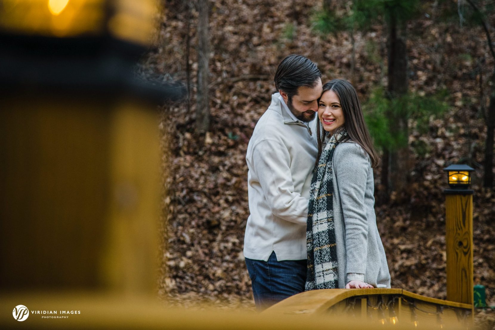 Romantic couple standing on lighted bridge over lake during Rocky Lake engagement session at dusk.